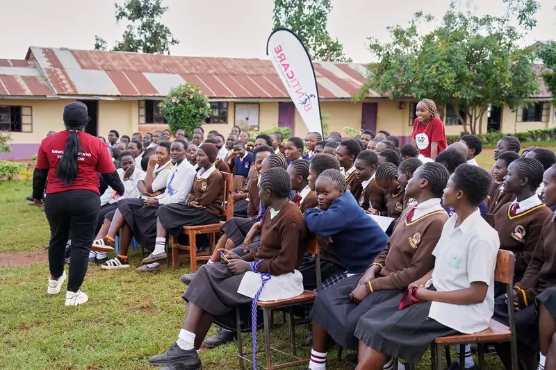 A trainer addressing a seated schoolgirl audience at an outdoor session beside a BintiCare banner